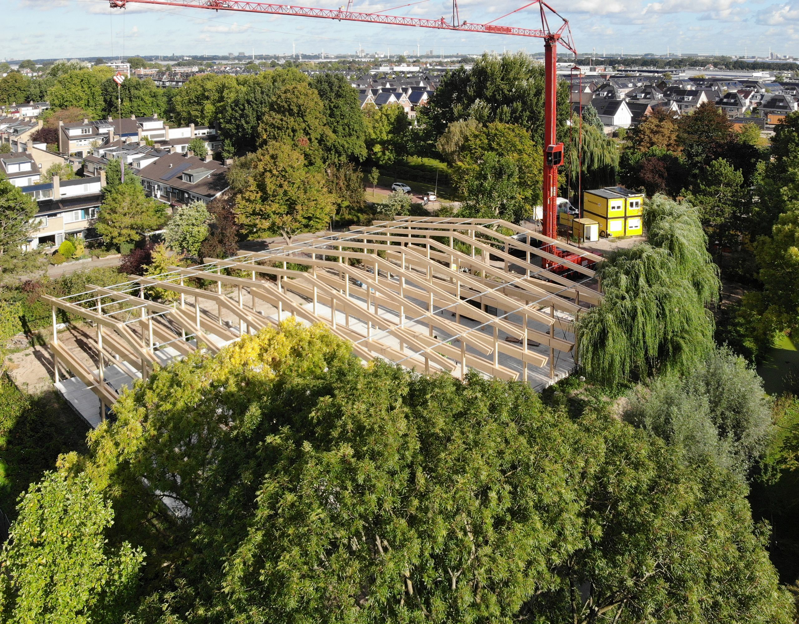 Dronebeeld van de houten draagconstructie van Kindcentrum De Wiekslag tijdens de bouw, tussen bestaande bomen